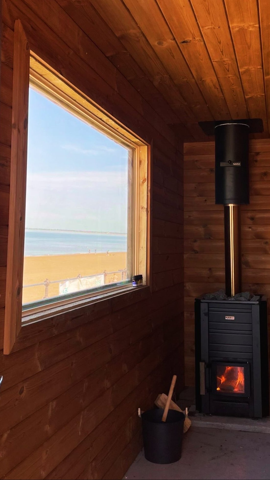Wooden cabin interior with a large window overlooking a beach and a wood stove.