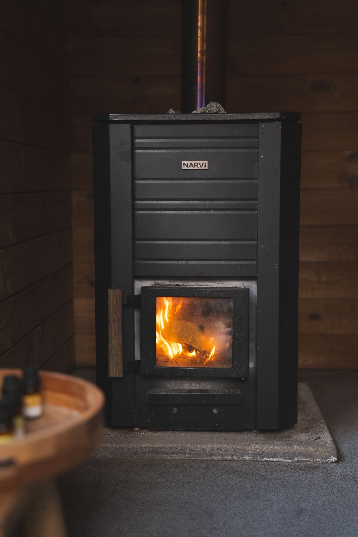Black wood stove with visible flames inside, set against a wooden wall.